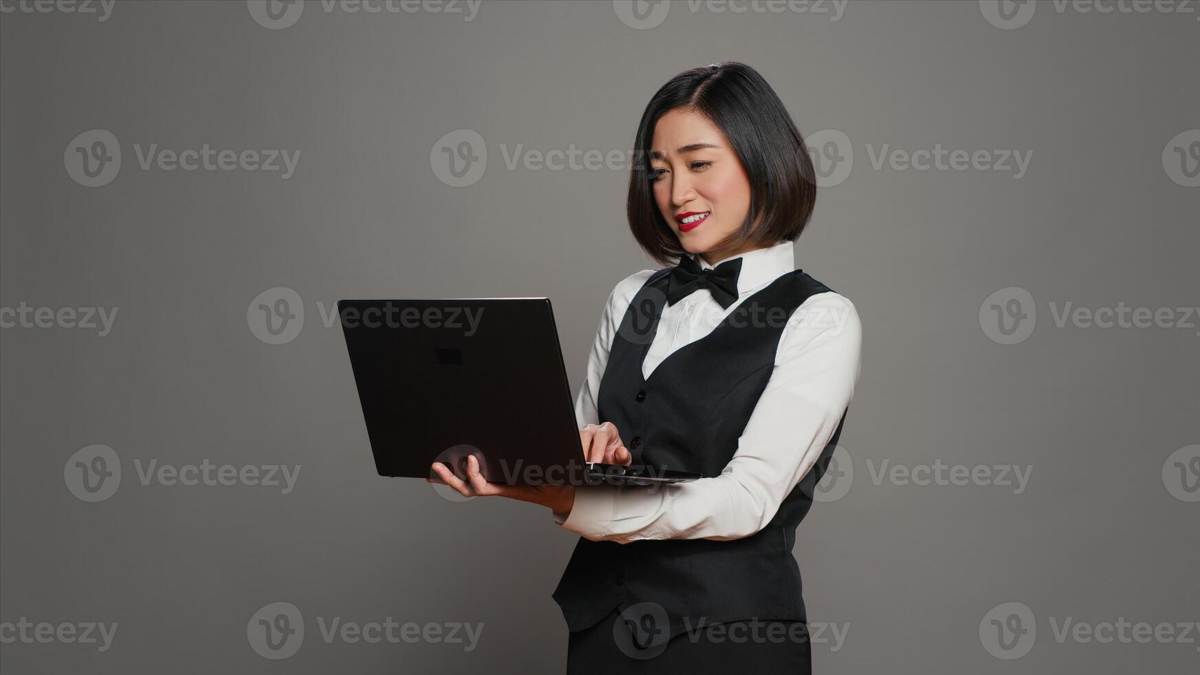 Asian hotel employee registering people using laptop at front desk, confirming online bookings and room reservations in studio. Receptionist posing with pc over grey background. Camera A. photo
