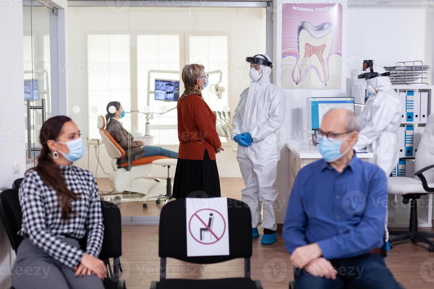 Senior woman patient in stomatology clinic discussing with doctor dressed in ppe suit as safety precaution during global pandemic with coronavirus. People talking in stomatology waiting room. photo