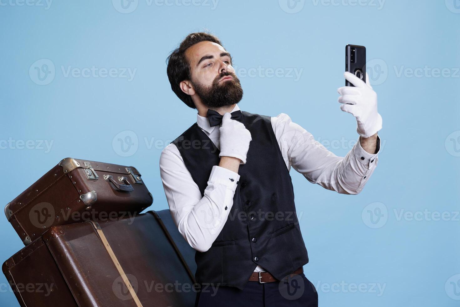 In studio, bellman in uniform takes pictures while using mobile device for selfies and acting foolish against blue background. Classy hotel porter does photos with his phone while being on duty.