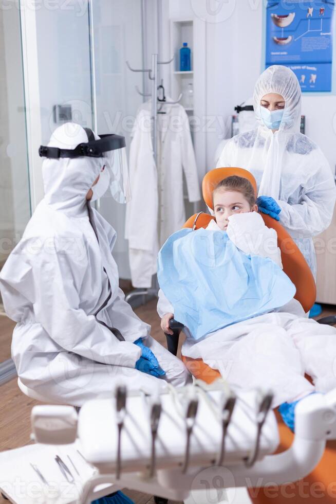 Little girl with painfull expression sitting on dental chair wearing bib. Stomatologist during covid19 wearing ppe suit doing teeth procedure of child sitting on chair. photo