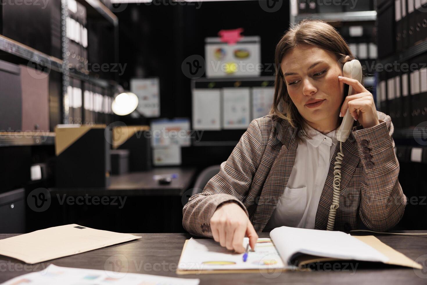 Bookkeeper having remote conversation, working overtime at administrative report in storage room. Caucasian businesswoman discussing accountancy folders, checking corporate documentation photo
