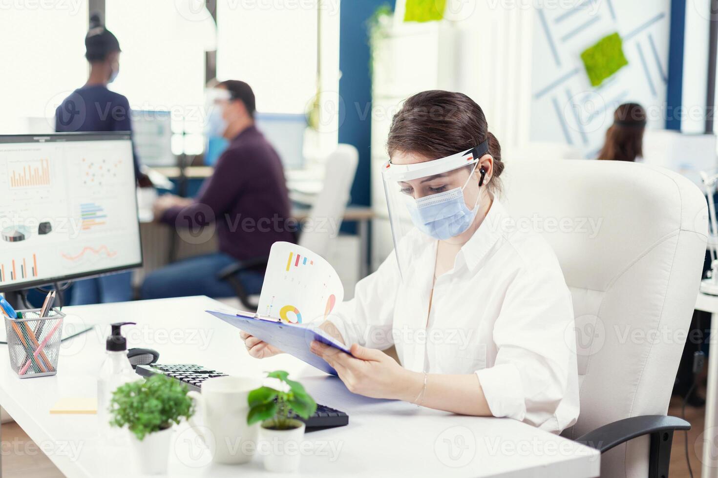 Business consultant on a call with consultant using wireless headphones during covid-19. Employees with visors working in corporate company workspace respecting social distance analysing data and graphs. photo