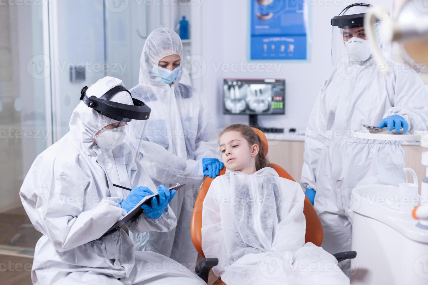 Dentist doctor with face mask and coverall holding clipboard dressed in coverall Stomatologist during covid19 wearing ppe suit doing teeth procedure of child sitting on chair. photo