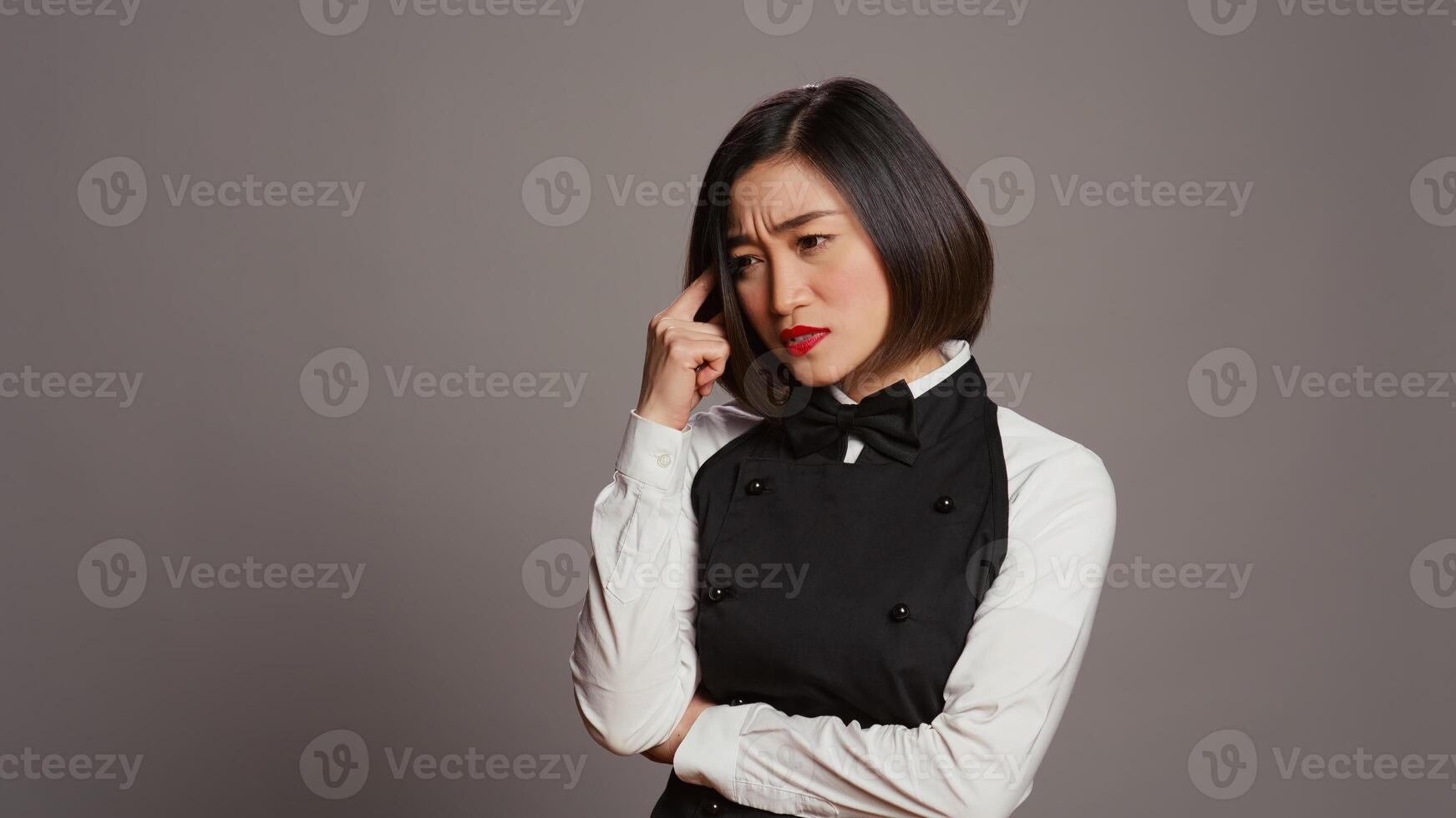 Restaurant hostess being thoughtful over grey background, thinking about new ideas to develop the menu for clients. Asian woman waitress feeling pensive, imagination in studio. Camera B. photo