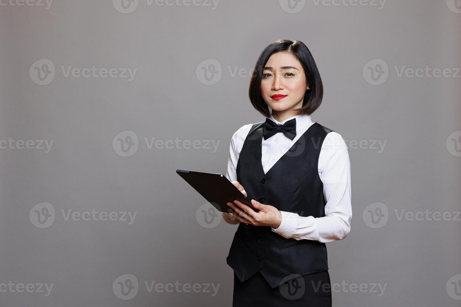 Smiling confident asian waitress wearing professional uniform, holding digital tablet and looking at camera. Young attractive hospitality industry receptionist using portable gadget portrait photo