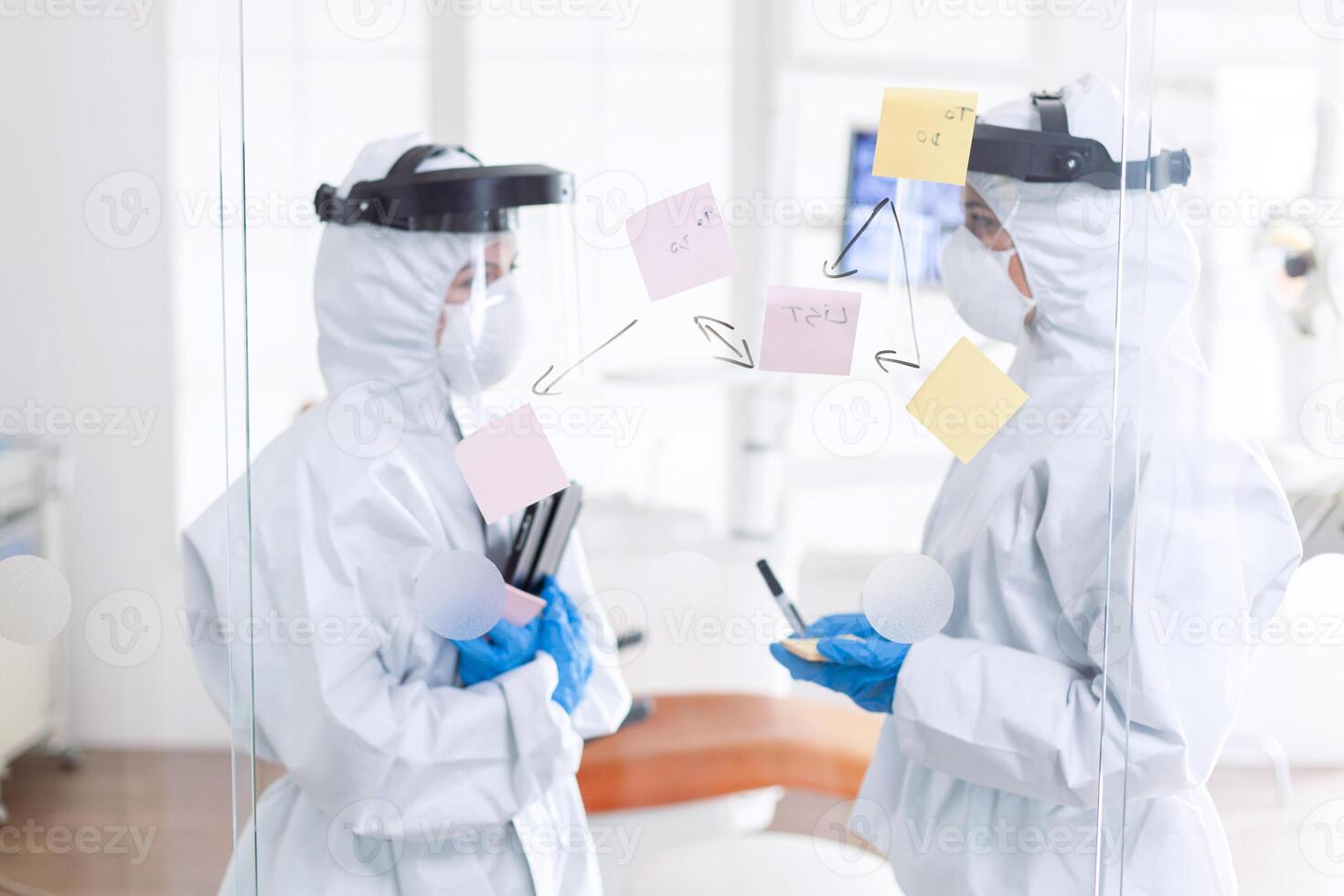 Tired dentist discussing about patient diagnosis wearing ppe suit. Medical team in stomatology office wearing coverall in dental office writing ideas on sticky notes. photo