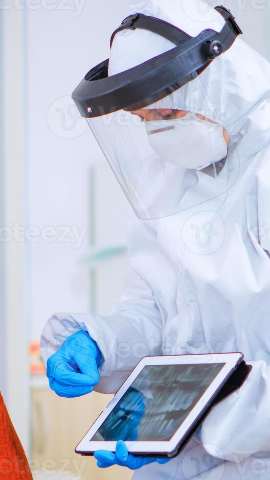 Woman sitting on chair in waiting area with protection mask listening doctor with overall looking on tablet in clinic with new normal. Assistant explaining dental problem during coronavirus pandemic photo