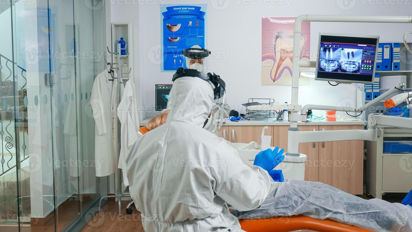 Orthodontist in protection suit explaining dental surgery to patient with toothache during covid-19 in stomatological office. Assistant and dentist doctor wearing coverall, face shield, mask, gloves photo
