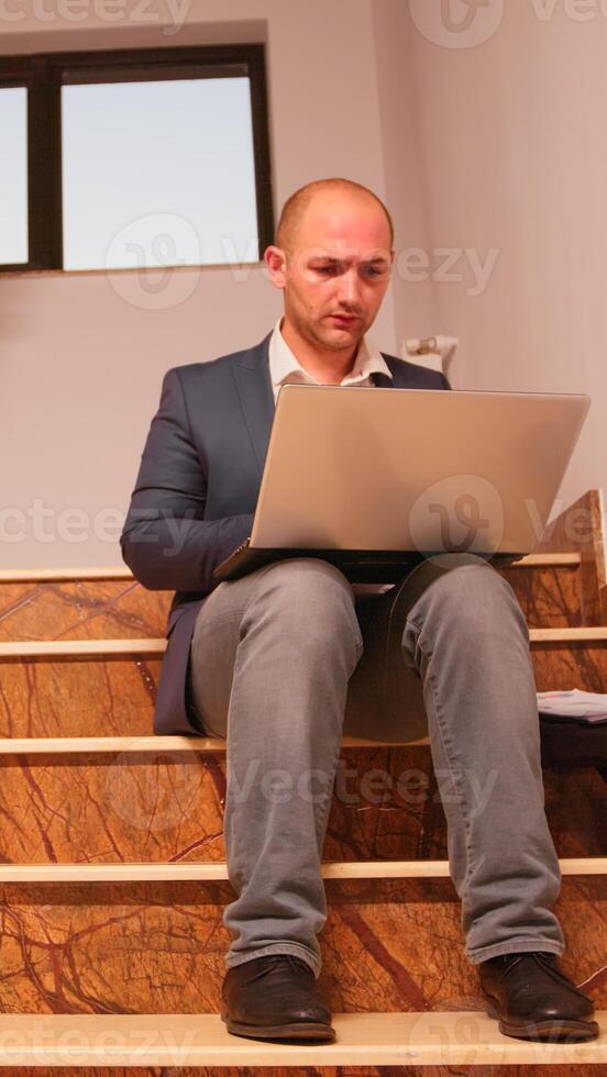 Overworked office executive sitting on stair typing on laptop looking on documents from clipboard while colleagues moving on staircase. Professional businesspeople working in modern financial building photo
