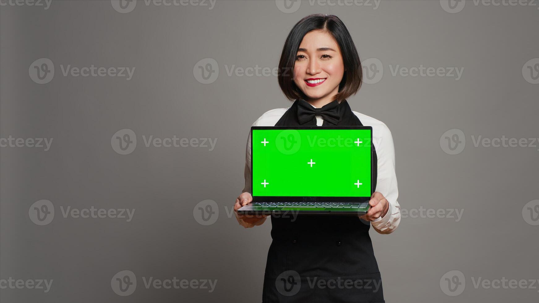 Asian server presenting greenscreen display on laptop in studio, wearing uniform with apron and bow. Restaurant hostess holding pc with blank mockup template, copyspace layout. Camera A. photo
