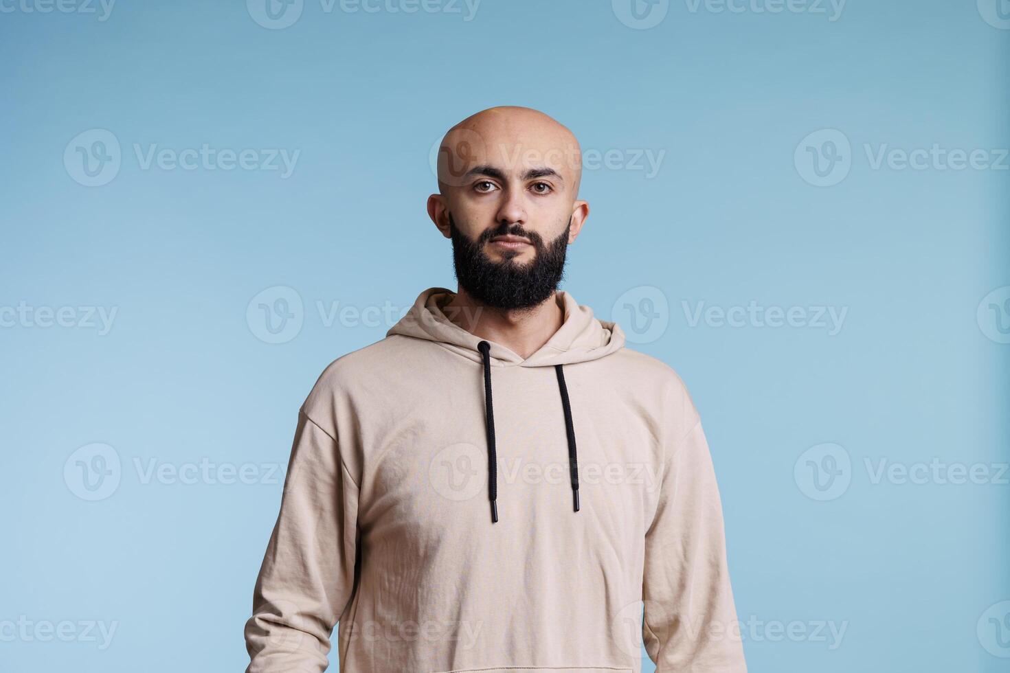 Young arab man standing in casual pose while looking at camera. Bald bearded person with neutral facial expression wearing casual beige hoodie clothes studio portrait on blue background photo