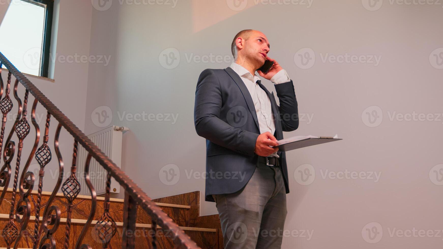 Furious businessman talking on smartphone standing on stairs in finance corporate company working overtime. Group of professional successful businesspeople working in modern finacial building. photo
