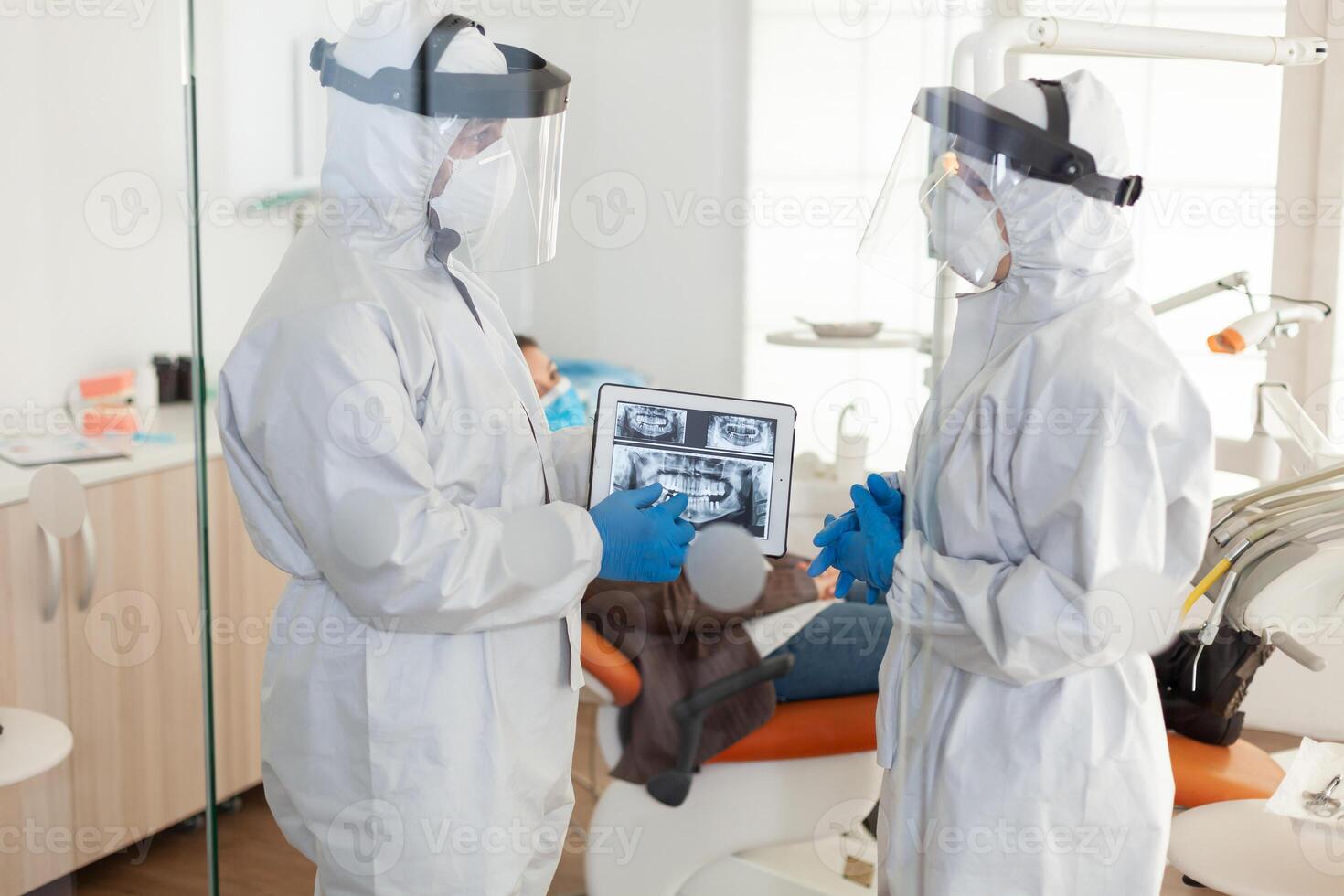 Dentist doctors with ppe suit analysing teeth x-ray using tablet in dental room, planning surgery during global pandemic while patient waiting on stomatological chair. photo