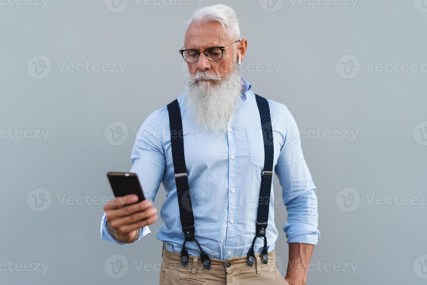 mayor hombre utilizando móvil teléfono inteligente y escuchando música con inalámbrico auriculares - Moda mayor masculino trabajando con tecnología dispositivos foto