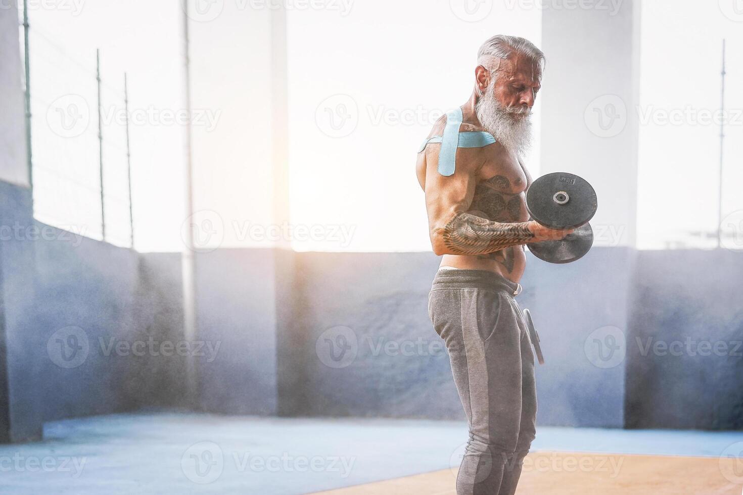 Fitness beard man doing biceps curl exercise  inside a gym - Tattoo senior man training with dumbbells in wellness club center - Body building and sport fit concept photo