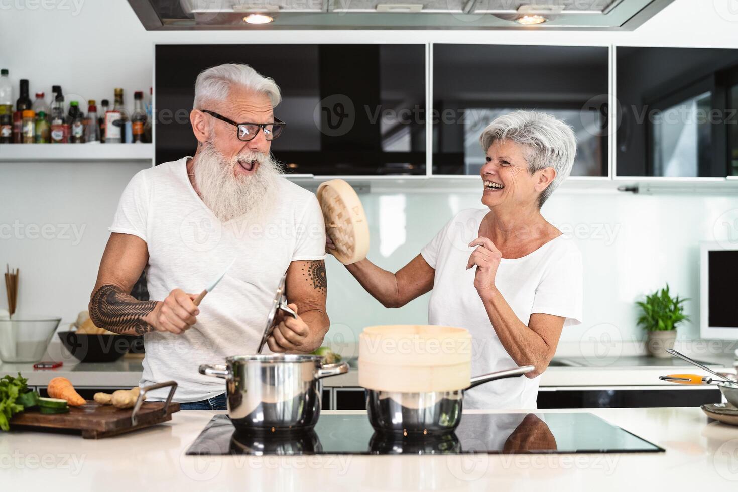 Happy senior couple having fun cooking together at home - Elderly people preparing health lunch in modern kitchen - Retired lifestyle family time and food nutrition concept photo