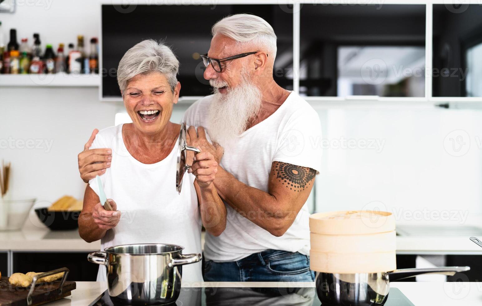 Happy senior couple having fun cooking together at home - Elderly people preparing health lunch in modern kitchen - Retired lifestyle family time and food nutrition concept photo