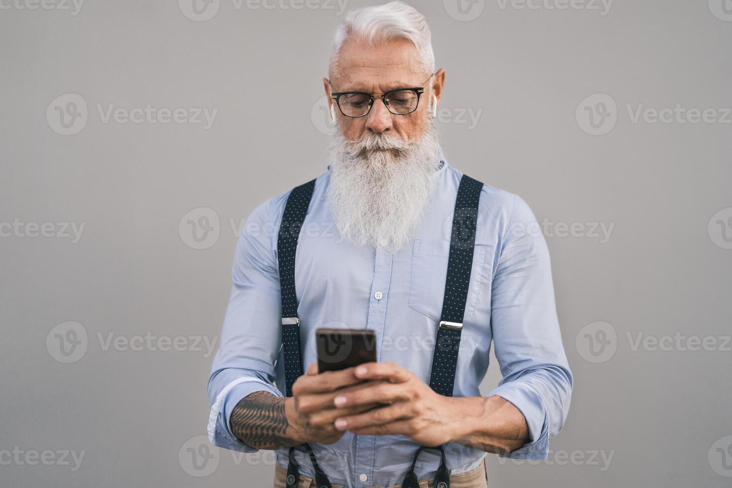 Senior man using mobile smartphone and listening music with airpods - Fashion bearded male working on digital marketing social media - Elderly lifestyle people and technology concept photo