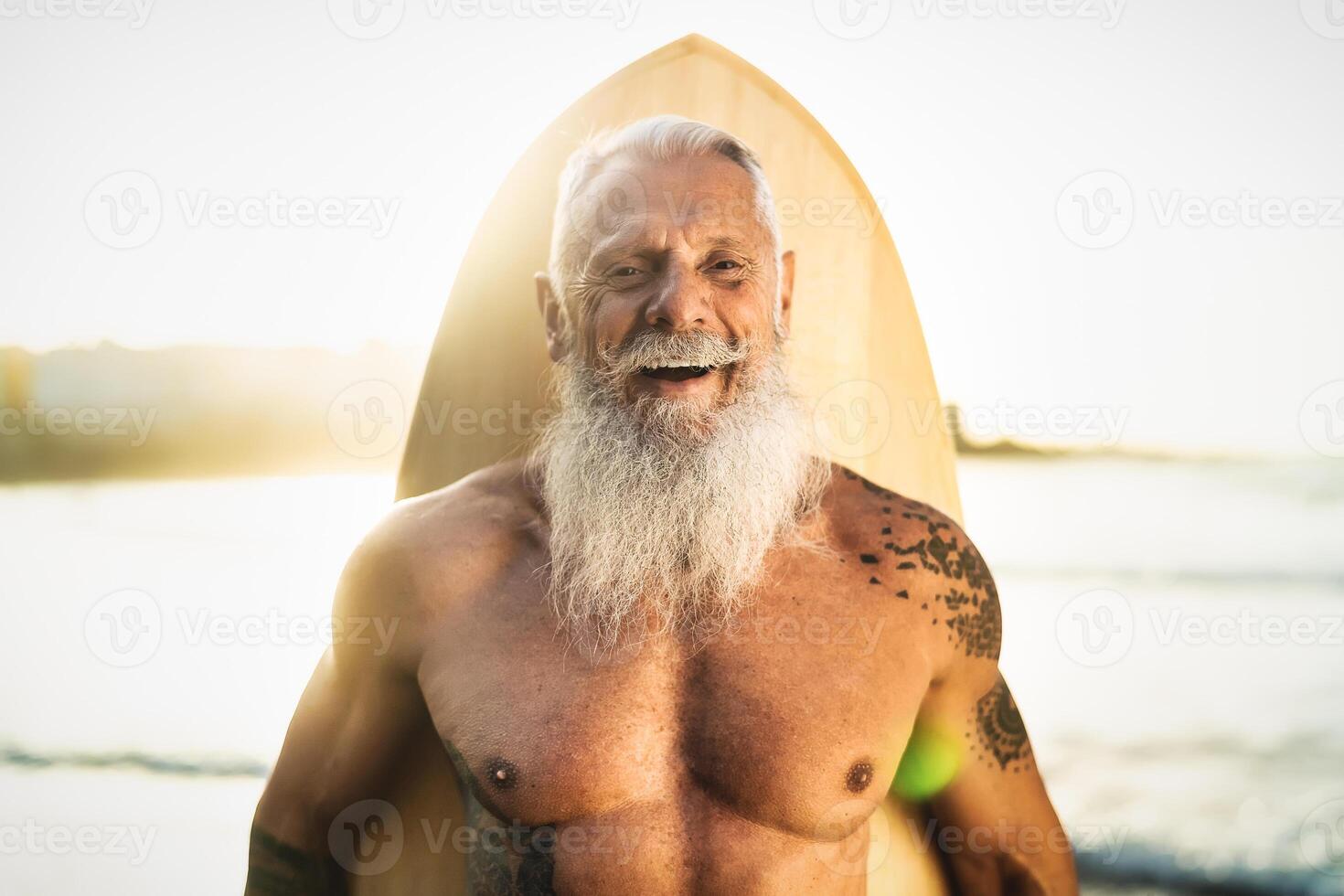 Fit senior male having fun surfing during sunset time - Retired man training with surfboard on the beach - Elderly healthy people lifestyle and extreme sport concept photo