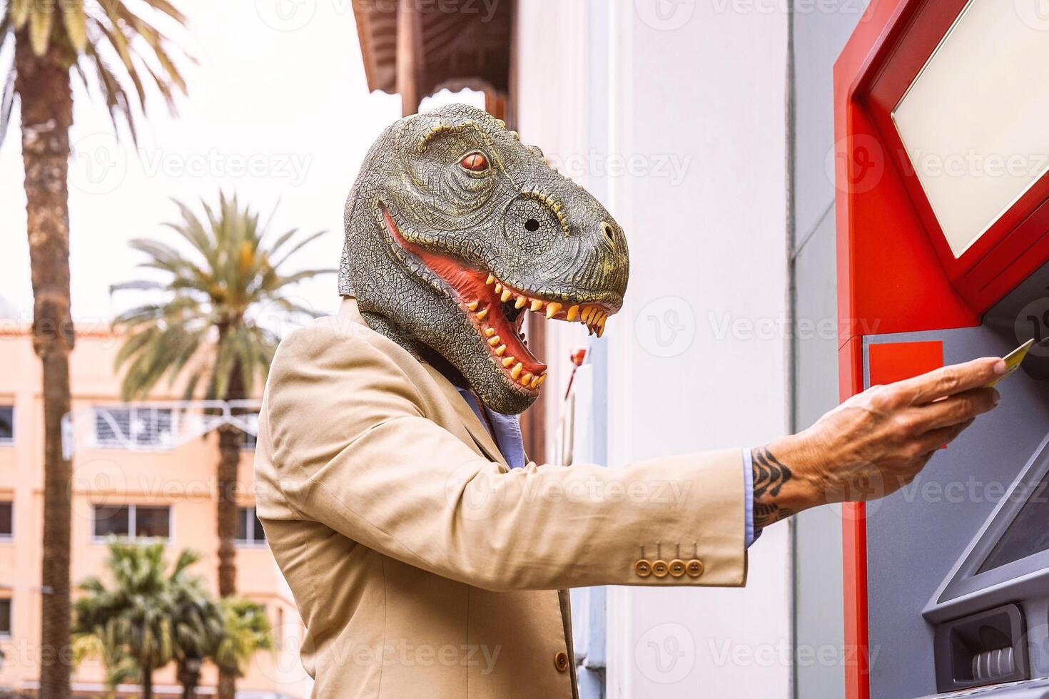 Senior man wearing t-rex dinosaur mask withdraw money from bank cash machine with debit card - Surreal image of half human and animal - Absurd and crazy concept of ATM advertise photo