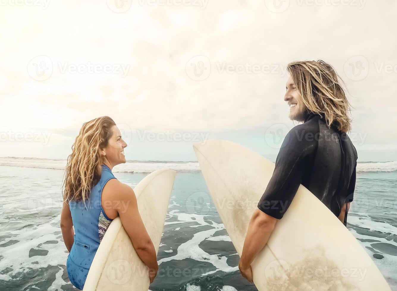 Surfers couple running together with surfboards on ocean at sunset time - Sporty fit friends having fun surfing on high waves - Travel and extreme sport culture lifestyle concept photo