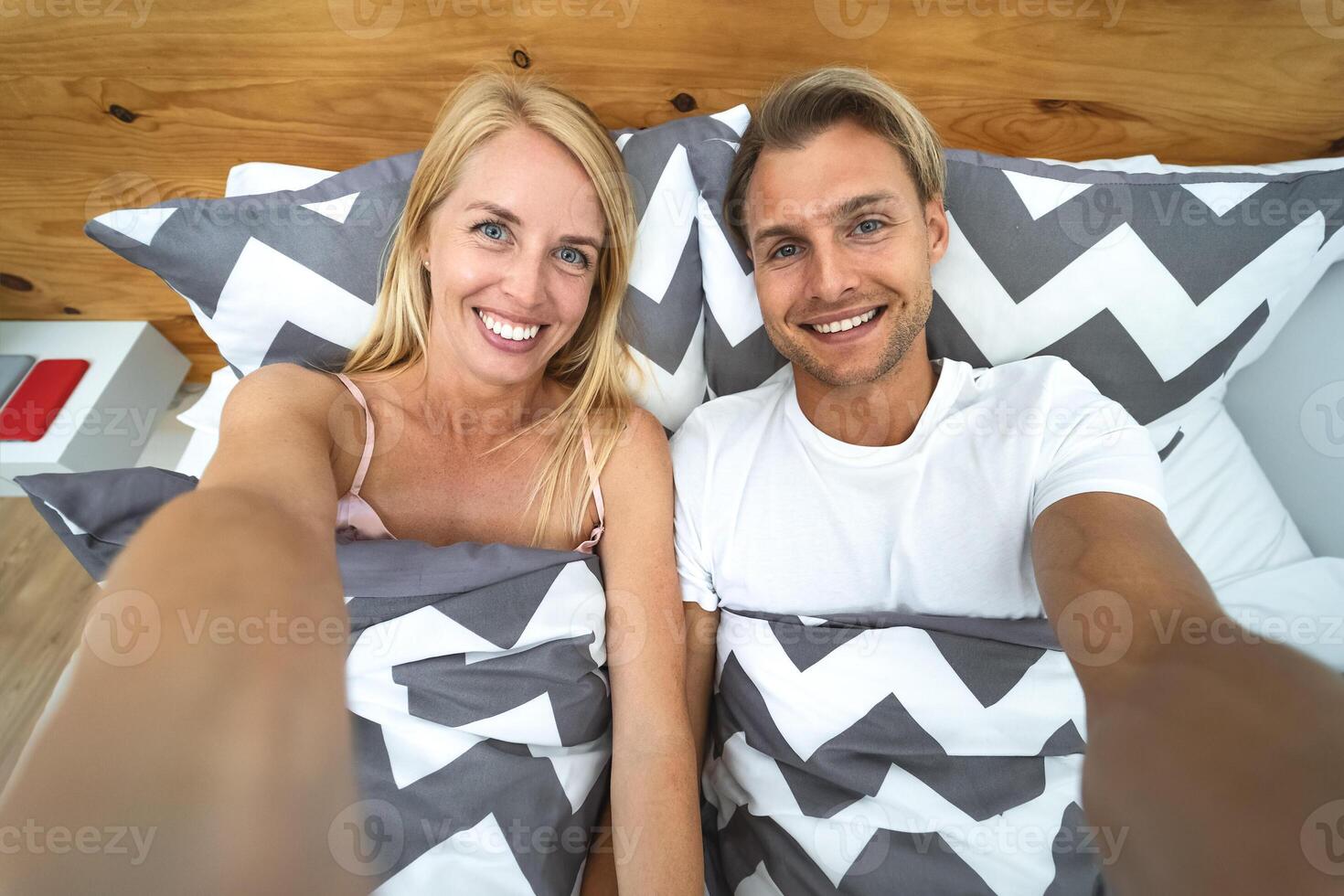 Young couple taking selfie in bed - Happy lovers having fun taking self photos lying on bed below blankets at home - Love relationship and youth culture lifestyle concept