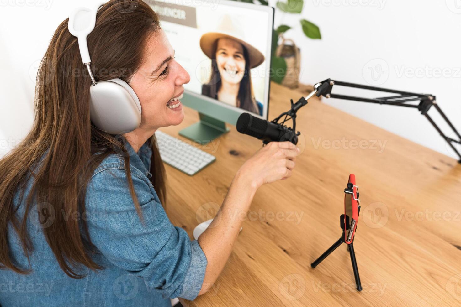 Mature woman recording a podcast using microphone and laptop while streaming on social network with mobile smartphone from her home studio photo