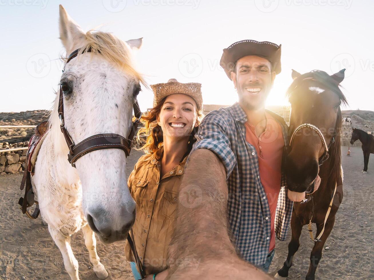 Happy couple taking selfie with horses inside stable - Young farmers having fun with animals in corral ranch - Human and animals relationship lifestyle concept photo