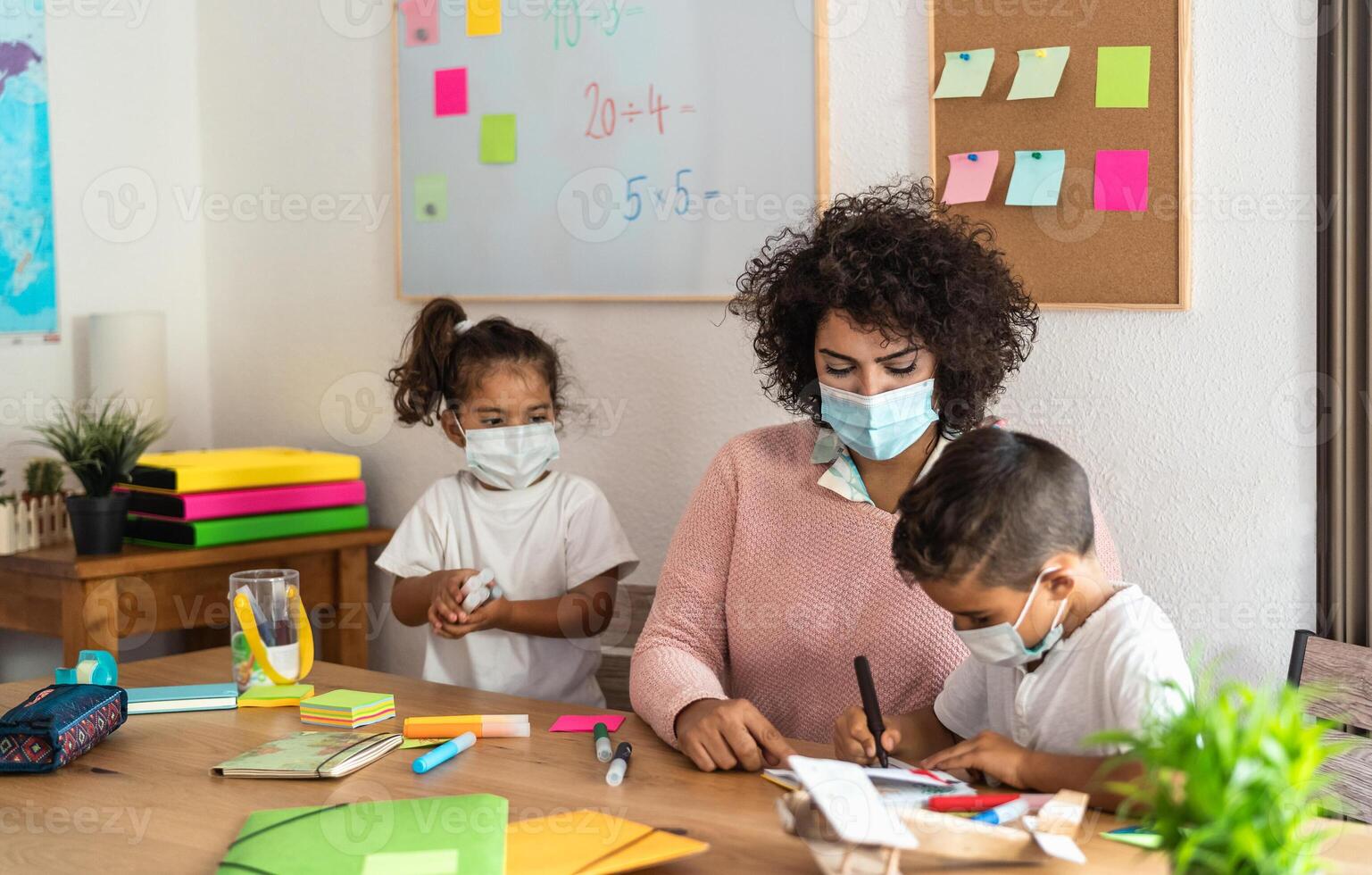 Female teacher with children drawing in preschool classroom while wearing face protective mask during corona virus pandemic - Healthcare and education concept photo