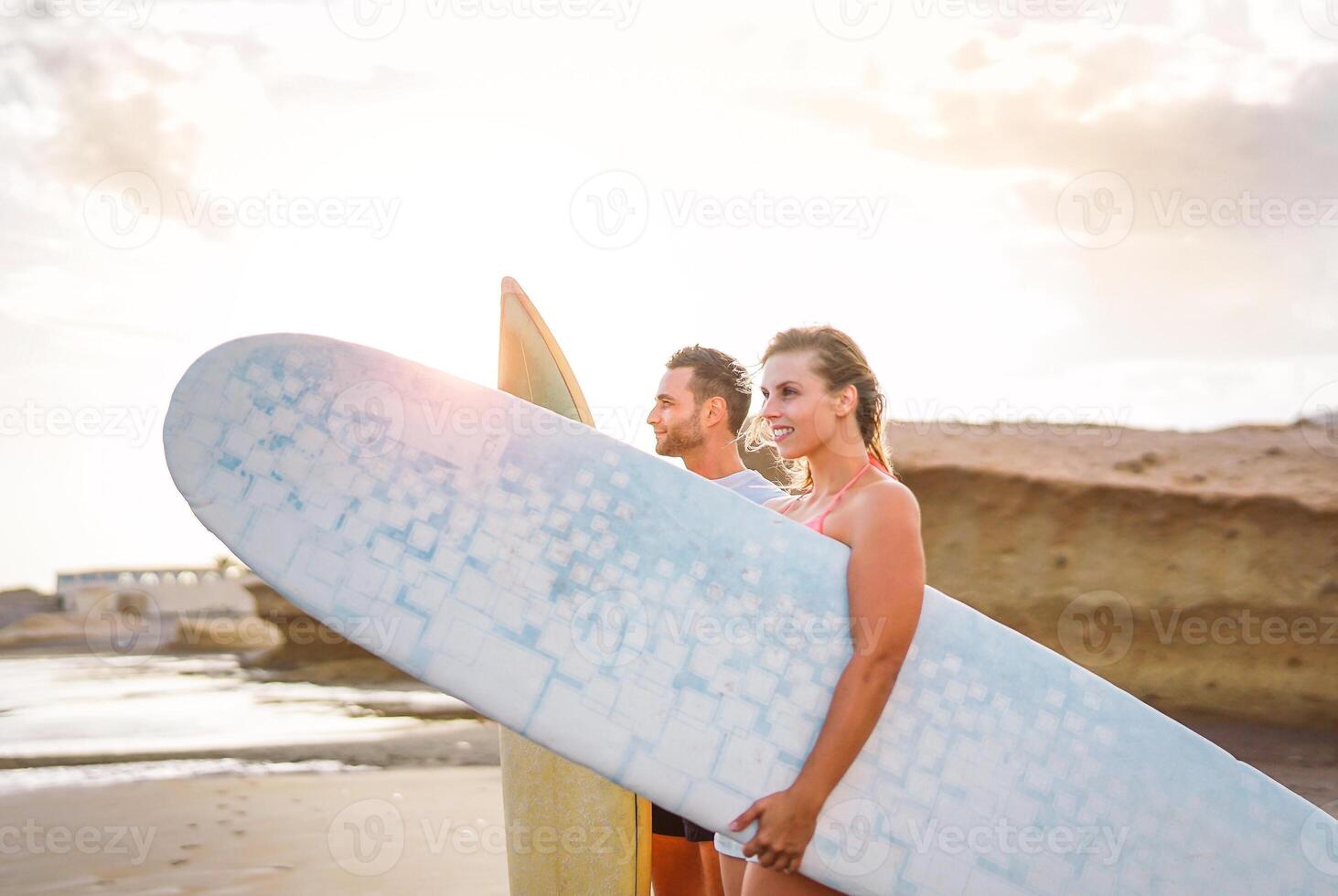 Young couple of happy surfers standing on the beach holding surfboards preparing to surf on high waves during a magnificent sunset - People, lifestyle, extreme sport concept - Focus on man photo