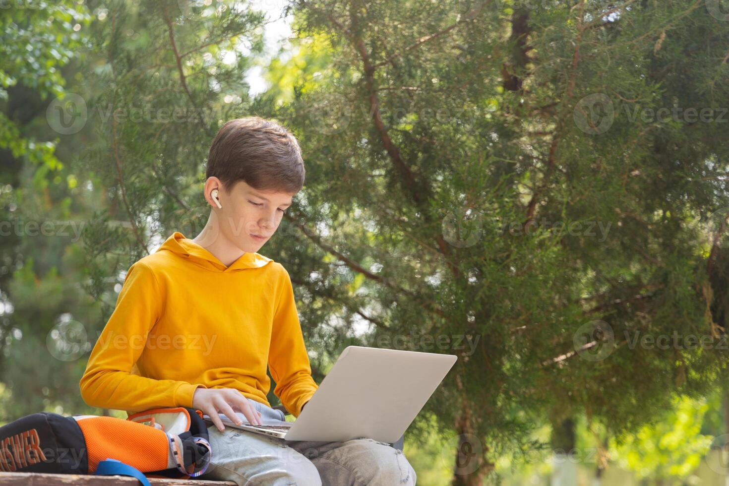 Thoughtful teenager boy resting. Holding and using a laptop for networking on a sunny day, outdoors. photo