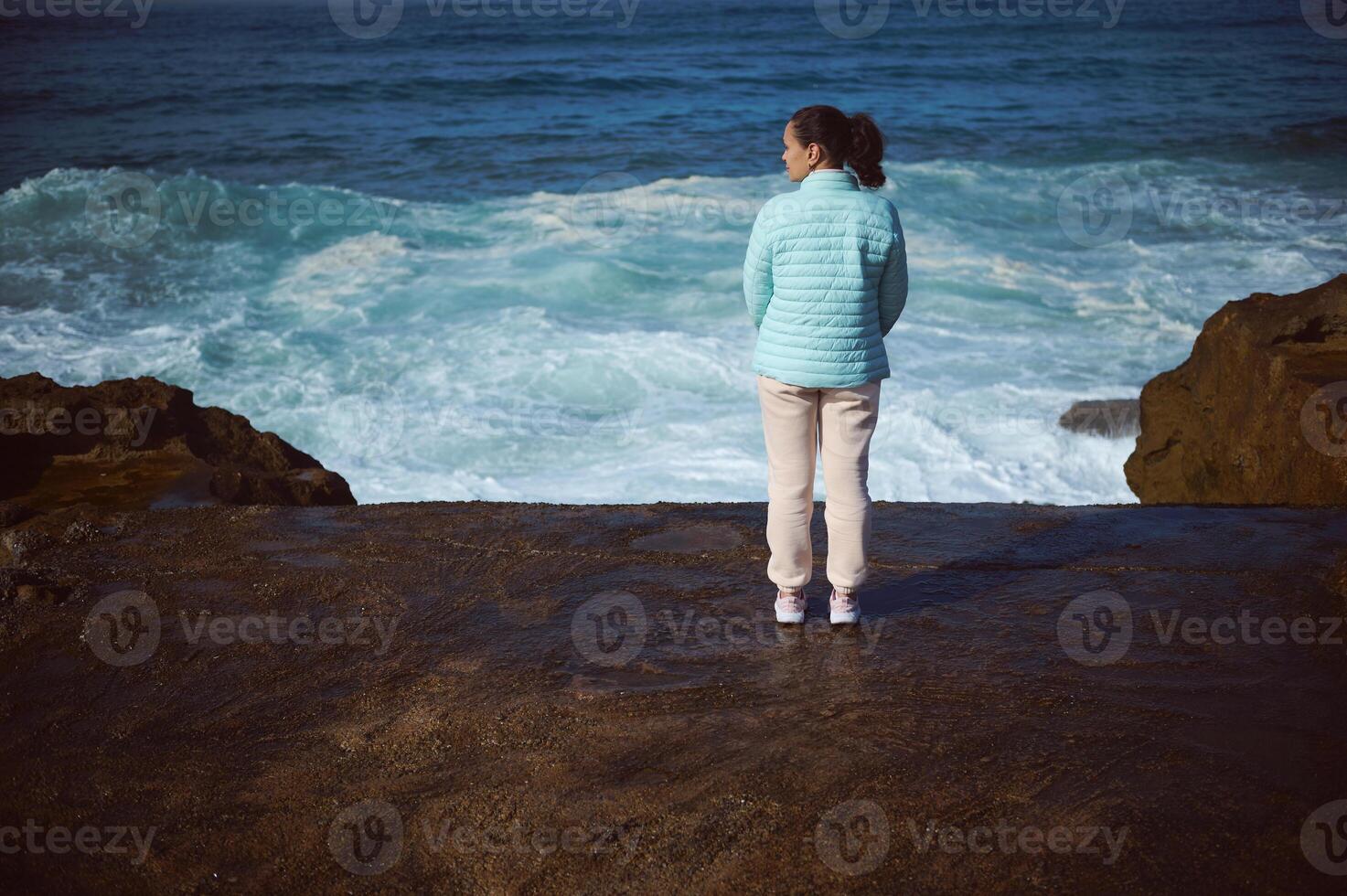 joven mujer disfruta conexión con naturaleza, en pie en rocoso acantilado, contemplando olas salpicaduras rotura en promontorio. foto