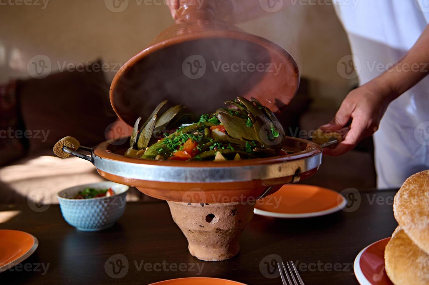 Close-up hands of a housewife opening the clay lid of Moroccan tagine while serving table for dinner at home photo