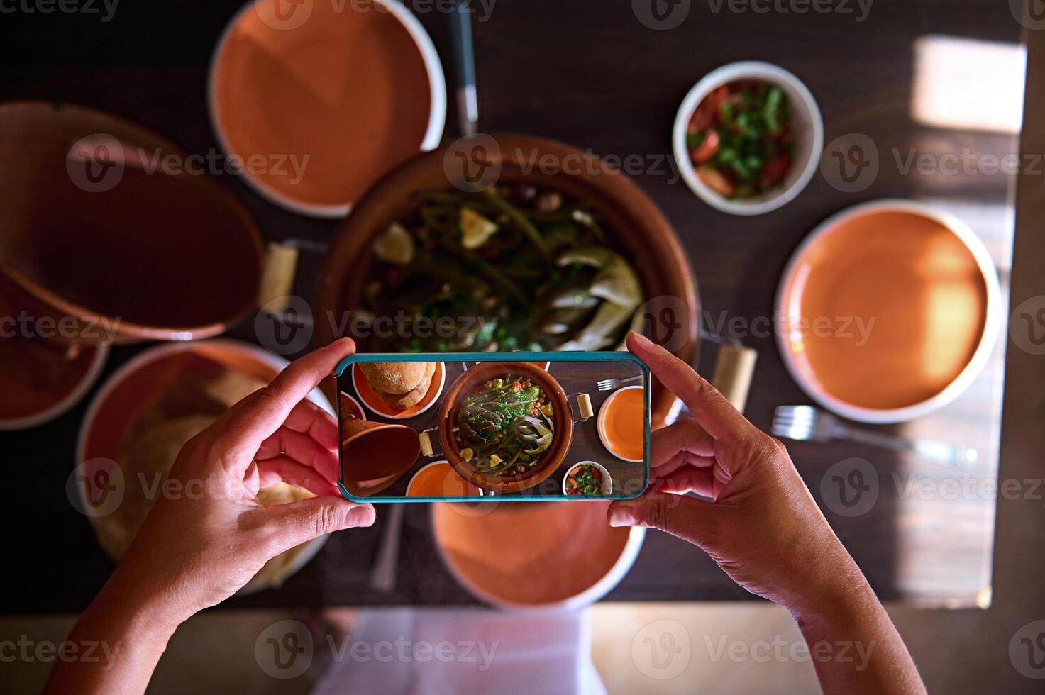 Directly above food blogger hands using smart mobile phone, taking photo of steamed veggies in clay dish tagine