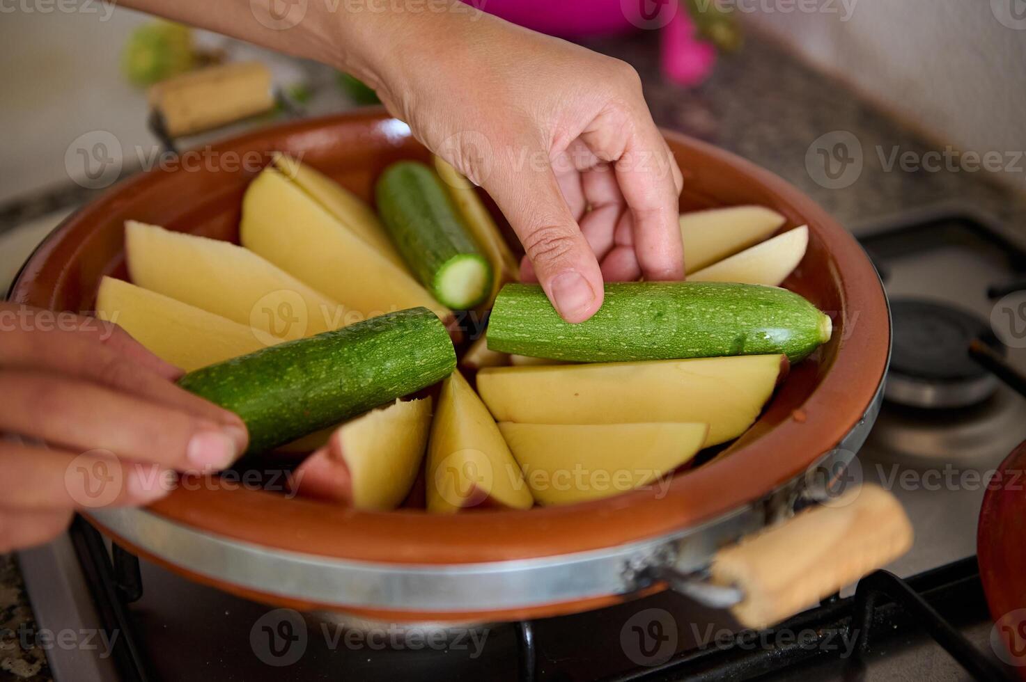 de cerca ama de casa manos poniendo calabacín y patata en el arcilla plato mientras Cocinando marroquí tagine en el cocina foto