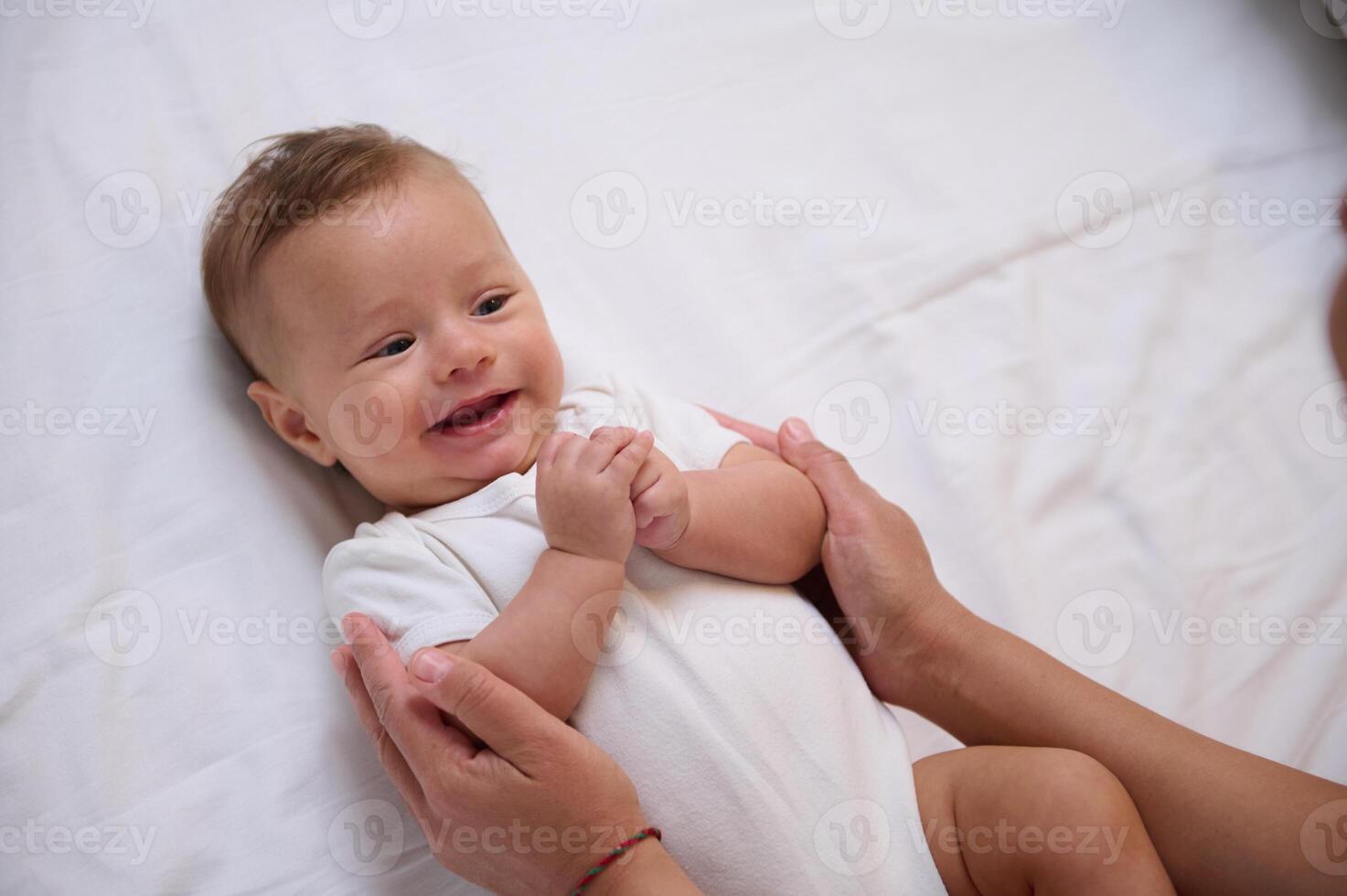 Adorable little baby boy in white bodysuit lying on the bed and connecting with his mother, smiling looking at her photo