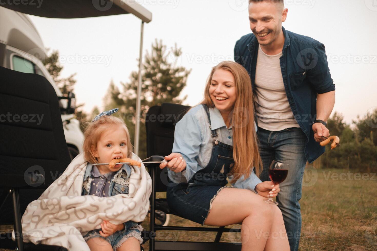 Mom feeds a sausage cooked near a motorhome on a campfire. Dad laughs watching photo