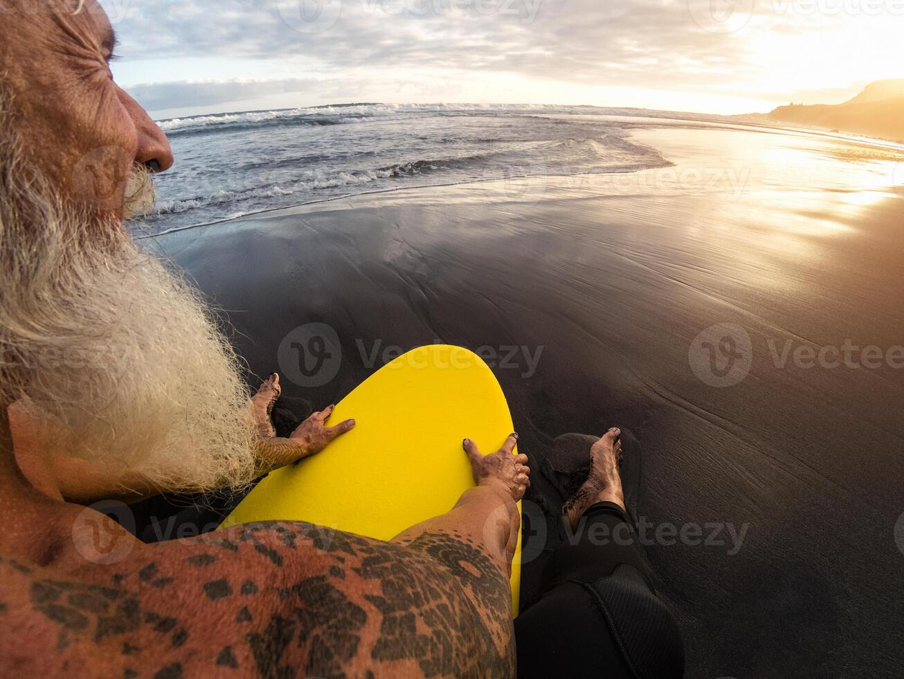 Happy fit senior man sitting on surfboard watching sunset time - Back view bearded surfer having fun on surfing day - Extreme sport and health people lifestyle concept photo