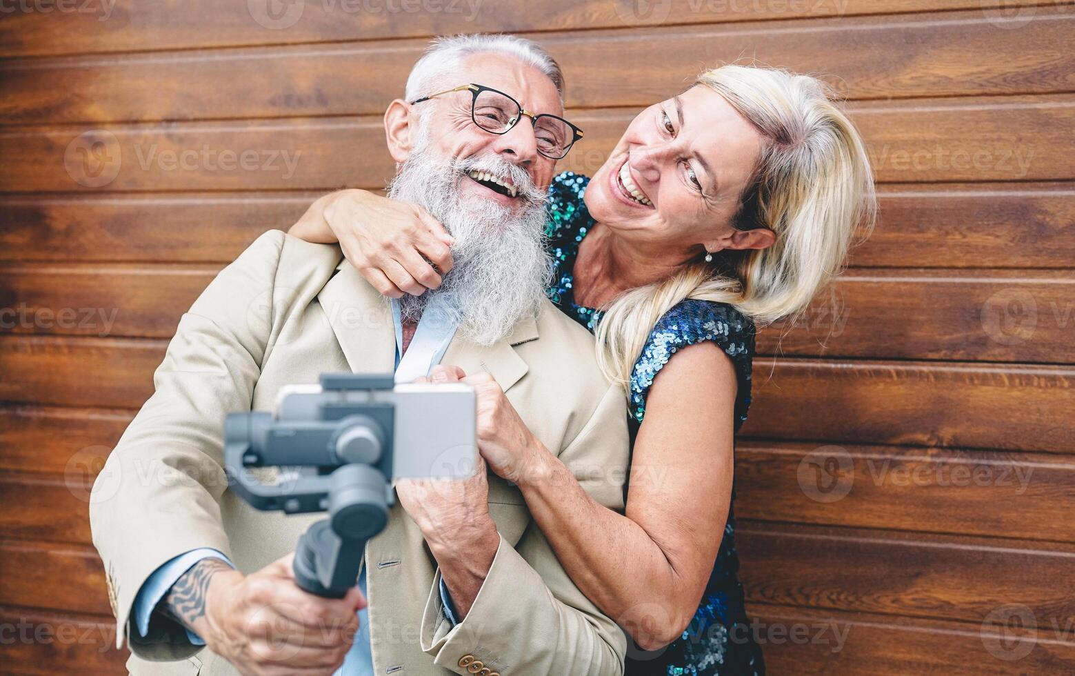 Happy seniors couple recording video with gimbal mobile phone outdoor - Mature fashion people having fun with new trends smartphone apps - wooden background - Elderly lifestyle and technology concept photo
