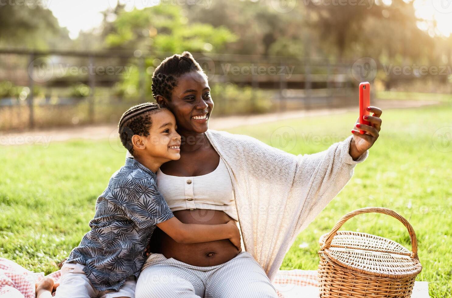 Happy African pregnant mother taking selfie with his son while doing a picnic during weekend in public park - Maternity and parents lifestyle concept photo