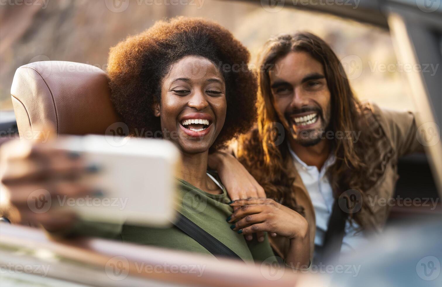 Happy young couple taking selfie with mobile smartphone while doing road trip - Travel people having fun driving in convertible car discovering new places - Relationship and vacation lifestyle concept photo