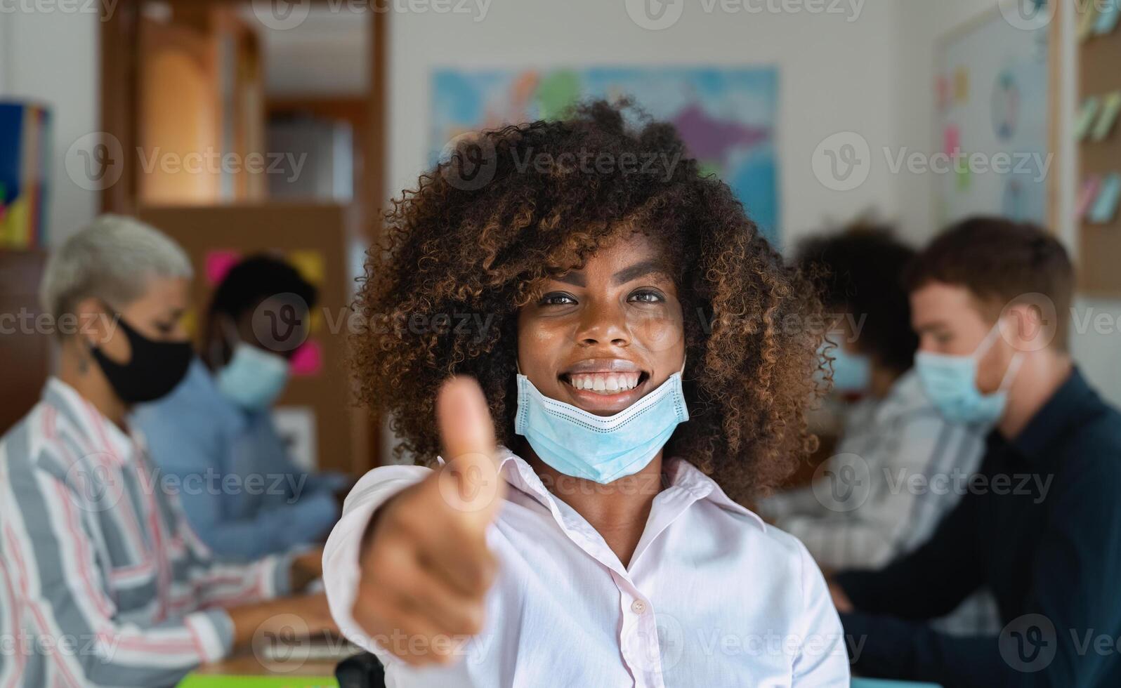 African woman thumb up wearing face mask in co-working creative space with team work - Young people working during corona virus pandemic photo