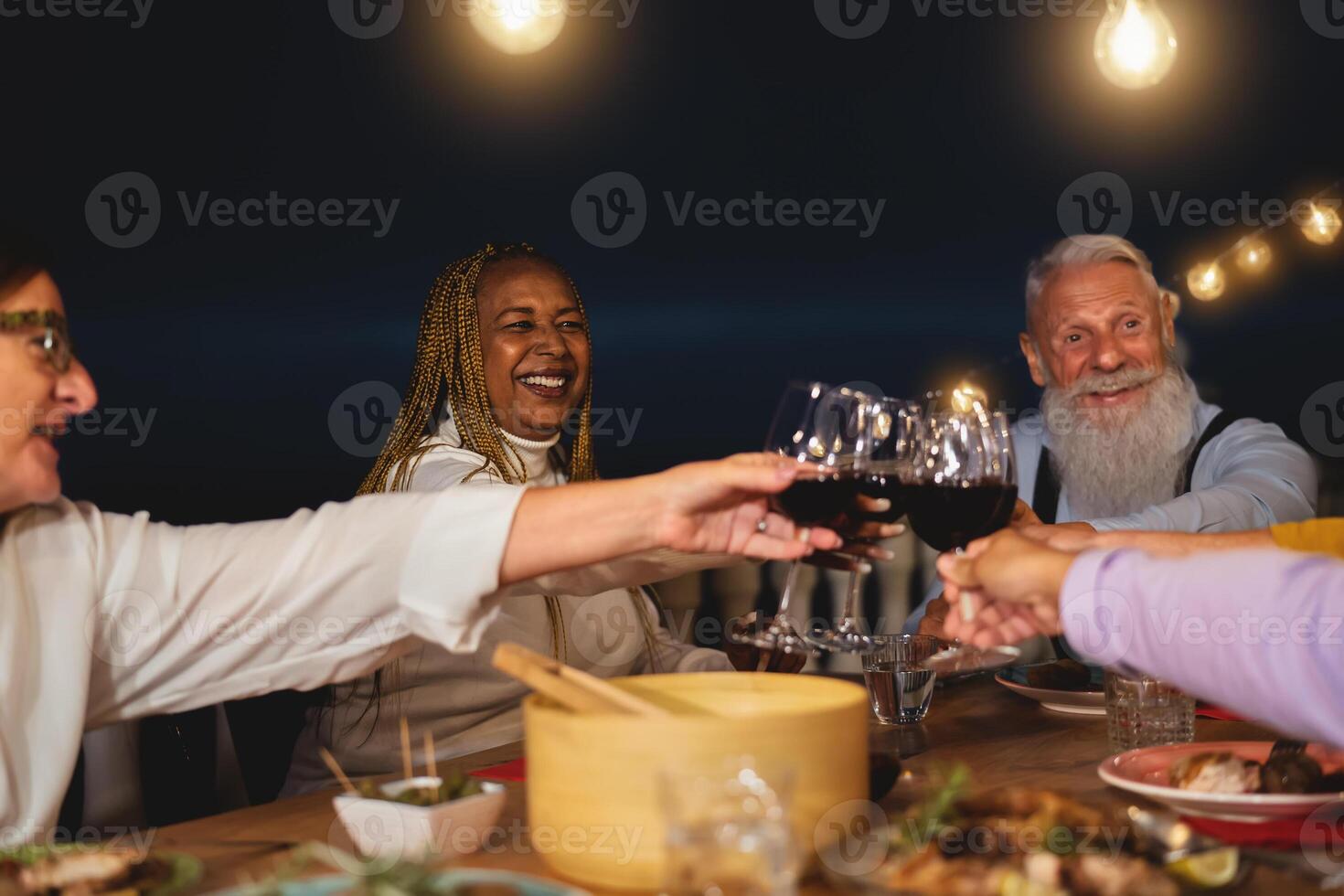 Happy multiracial senior friends toasting with red wine glasses together on house patio dinner - Elderly lifestyle people and food concept photo