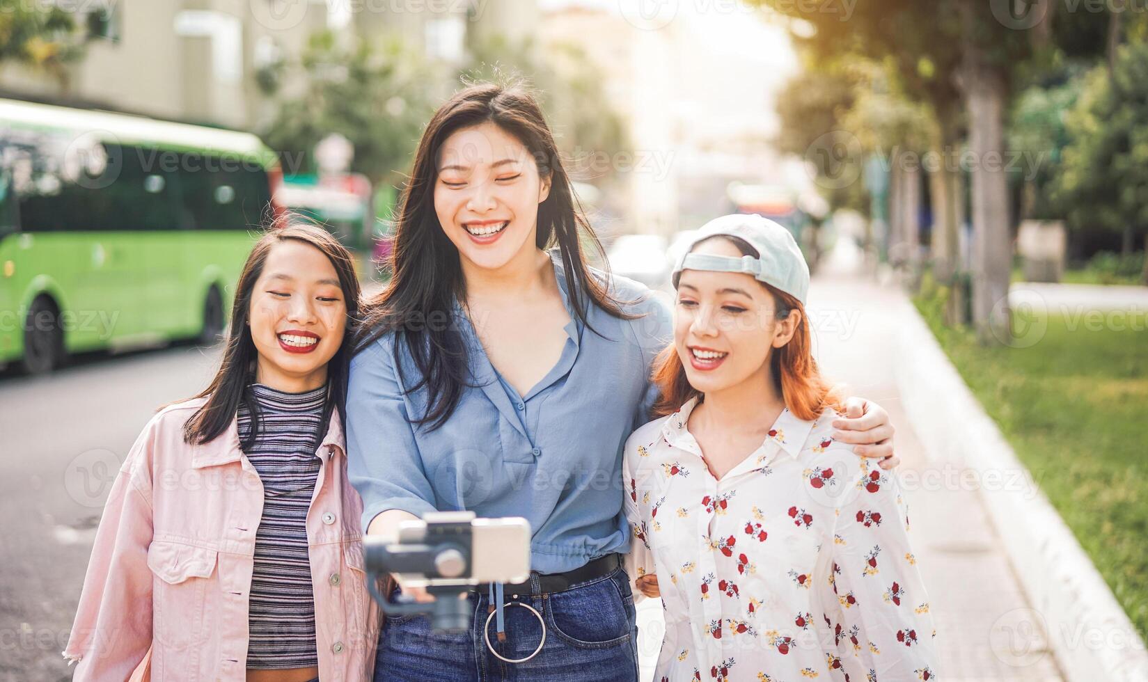 Happy Asian friends vlogging in city bus station - Trendy young people using gimbal smartphone outdoor - Friendship, technology, youth lifestyle and social media concept photo