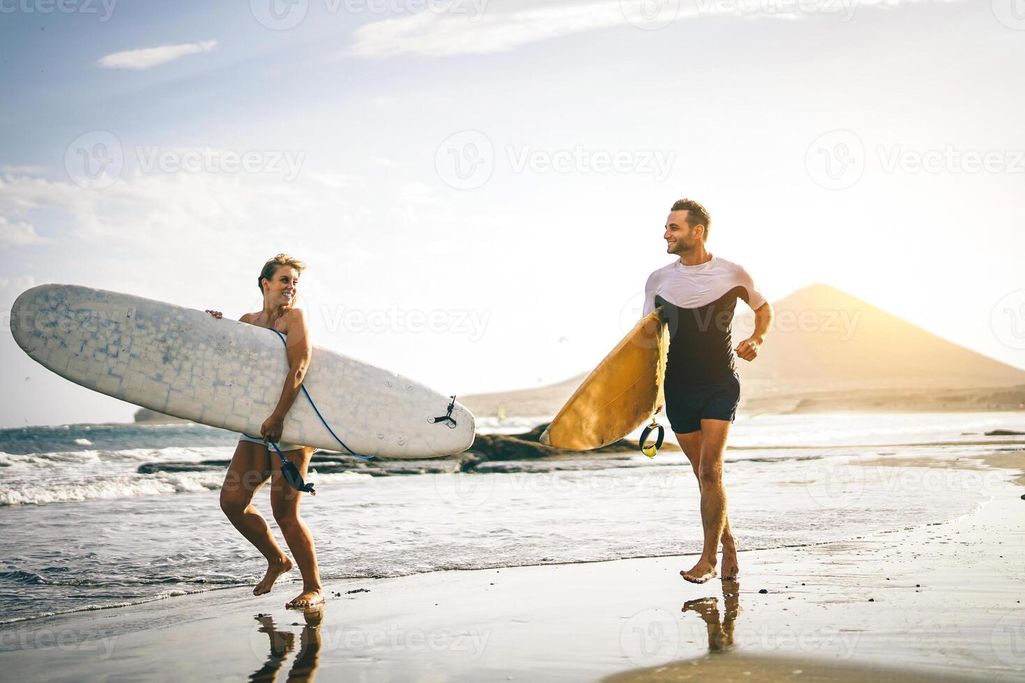 Young couple of surfers running with surfboards on the beach at sunset - Happy lovers going to surf together - People, sport and lifestyle concept - Vintage filter - Focus on woman board photo