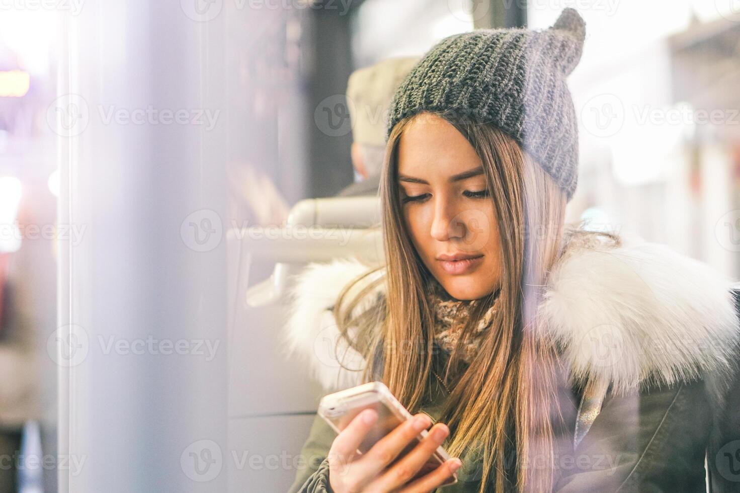 Young woman using her smart phone while sitting in a bus - Pretty girl connecting on her social network with her mobile phone - Concept of youth people addiction to new technology photo