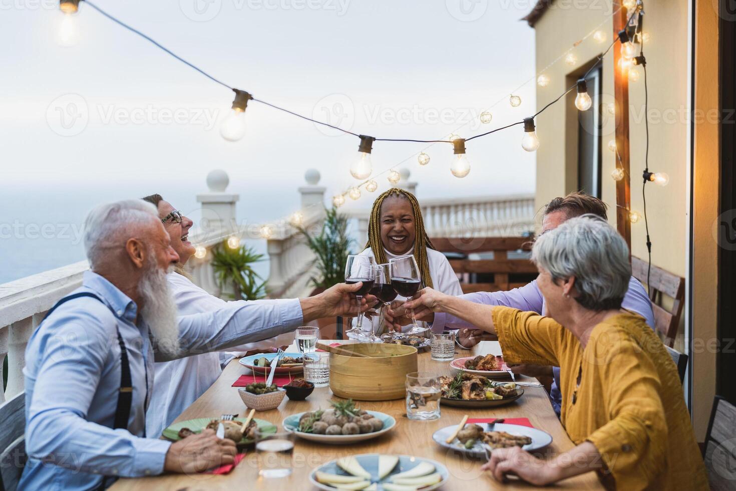 Happy multiracial seniors toasting with red wine glasses together on house patio dinner - Elderly lifestyle people concept photo