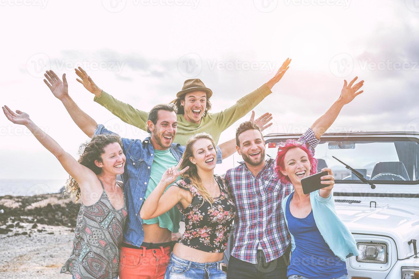 Group of happy friends taking a selfie using mobile smart phone during a road trip with jeep car - Travel people having fun spreading hands up while taking self photos in desert - wanderlust concept