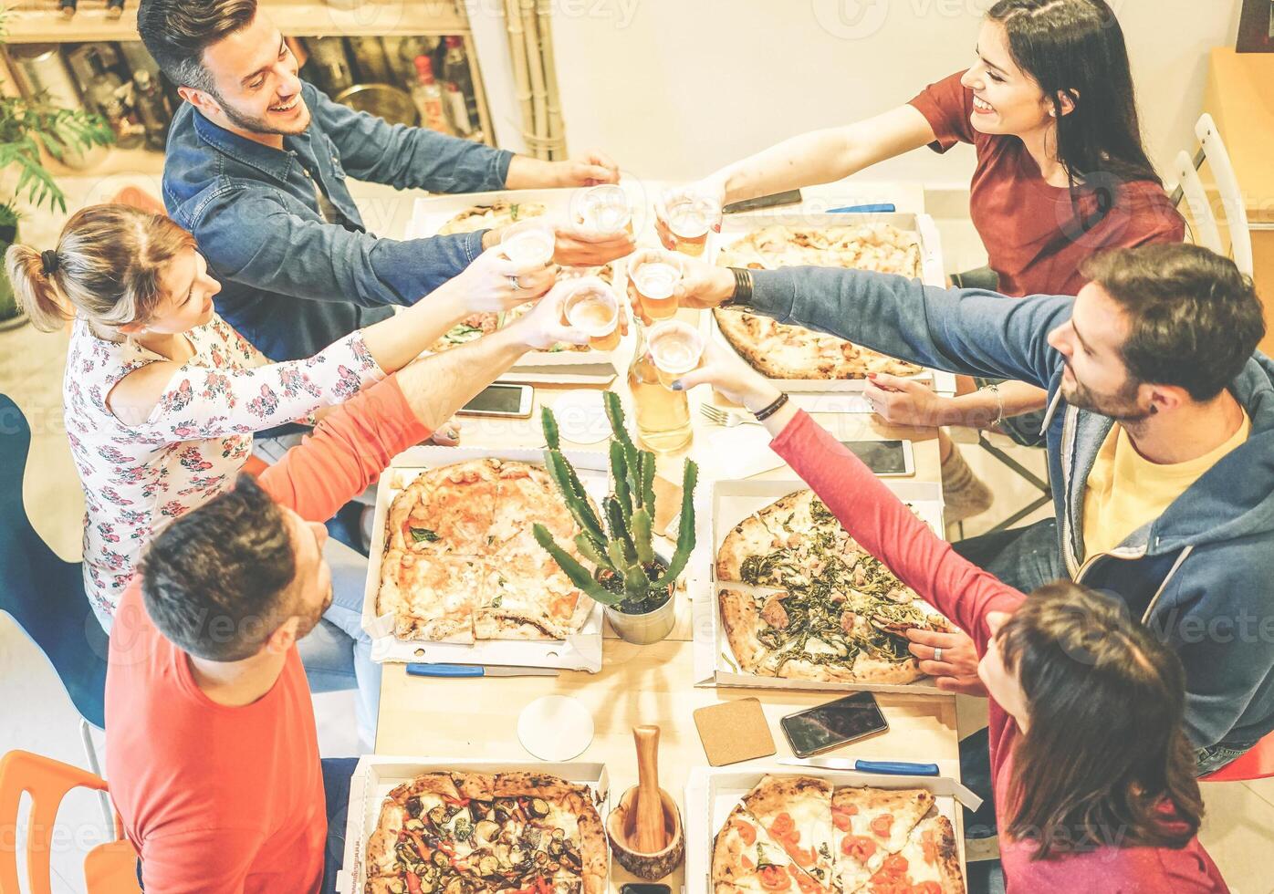 Group of friends toasting glasses of beer while eating pizza in their house - Happy people enjoying dinner together with tasty take away food at home - Concept of friendship, company, lifestyle photo