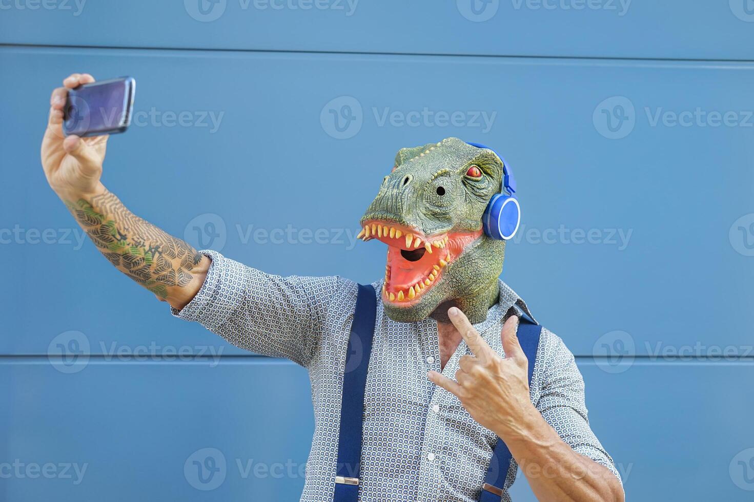 loco mayor hombre vistiendo tirano saurio Rex máscara mientras tomando selfie con móvil teléfono inteligente - hipster masculino teniendo divertido escuchando música y bailando al aire libre - absurdo surrealista y tecnología concepto foto